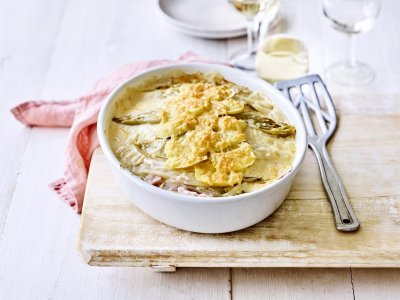 Er staat een ovenschotel op een houten plank, op de tafel. Er ligt een lepel naar de ovenschotel die gevuld is met een lasagne van witloof, ham, kaas en aardappelen. Er ligt ook een coraalroze servette bij.