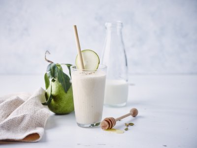 Een milkshake op basis van karnemelk, peer en banaan. Hij is geschonken in een hoog glas met rietje en stukje peer. Links van het glas ligt een verse peer en linnen servette. Rechts ligt de honinglepel op tafel.