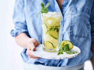 Een heerlijk frisse mocktail van Jupiler 0.0 en peach ice tea. Geserveerd met frisse limoenschijfjes in het glas. De vrouw draagt een blauw jeanshemd en staat mee in beeld.