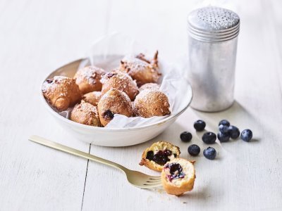 Oliebollen met blauwe bessen In een diep wit bord ligt een wit boterpapiertje met hierin enkele oliebollen. Er is eentje uitgeprikt en ligt op tafel doormidden gesneden, hier zie je duidelijk de blauwe bessen in zitten. Er is bloemsuiker over de oliebollen gestrooid voor een extra zoete toets.