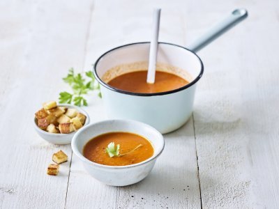 Op een houten witte tafel staat een kopje paprikasoep. De soepkom staat ook in beeld, dit is een lichtblauwe pot die mooi contrast geeft met de oranje-rode soep. De soep is afgewerkt met verse peterselie en croutons.