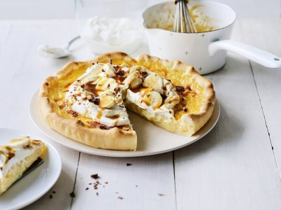 Rijsttaart met slagroom, banaan en gezouten karamel Op een witte houten tafel staat een klaargemaakte rijsttaart, hij is afgewerkt met vers opgeklopte slagroom, stukjes banaan en vloeibare karamel. Er is al een stukje aangesneden. Verder uit beeld staat het steelpannetje waarin de karamelsaus is klaargemaakt.
