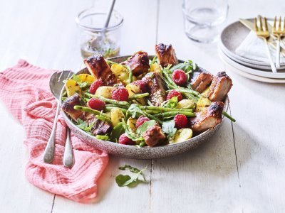 Salade van krieltjes, boontjes, frambozen en ribbetjes Een grote serveerschaal is gevuld met de aardappelsalade. Je ziet de framboosjes, boontjes en ribbetjes in de salade zitten. Er staat een torentje bordjes klaar met bestek om de salade te verdelen en op te eten.
