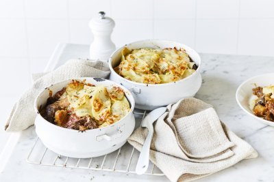 Twee witte stoofpotjes sttaan op een witte houten tafel en zijn gevuld met traag gegaard rundsvlees. De stoofpot is afgedekt met een laagje van aardappelschijfjes. De gratin is mooi goudgeel gebakken. Er staat een pepermolen langsheen de kookpotten, servette en grote lepel.