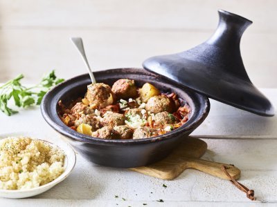 Tajine met gehaktballetjes van lamsvlees Zwartkleurige tajine met gehaktballetjes van lamsvlees, afgewerkt met verbrokkelde feta. Er staat een portie couscous naast de tajine.