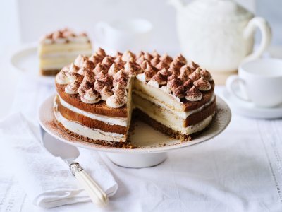Tiramisutaart met hazelnootbodem Op tafel staat een prachtige taart, het is een twist op de klassieke tiramisu. Hij staat op een witte taartschaal, en is afgewerkt met toefjes mascarpone en cacaopoeder. In de achtergrond staat een koffiekan en tasjes koffie.