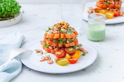 Tomaattorentje met grijze garnalen Een bord met daarop drie laagjes gesneden coeur de boeuf. Daarachter staat nog een bord met een potje met dressing en tuinkers.