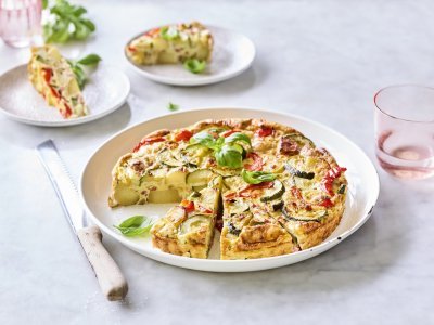 Tortilla met courgette, paprika en ham Een heerlijke aardappeltortilla ligt aangesneden op een groot wit bord. Er werden al twee stukjes uitgesneden en liggen geserveerd op kleine bordjes en afgewerkt met basilicum. Er staan licht gekleurd roze glazen langs het gerecht om de foto nog meer kleur te geven.