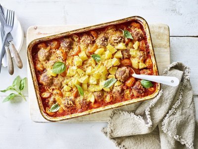 Traybake met gehaktballetjes in tomatensaus en krokante aardappelblokjes Op tafel staat een grote rechthoekige ovenschaal op een houten plank. Er staan witte bordjes langs met bestek om op te scheppen met een witte lepel. Bij de ovenschotel ligt ook een bruin servette. Het gerecht is afgewerkt met verse basilicum die mooi contrast geven op de tomatensaus en goudgele krokante aardappeltjes.