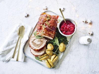 Varkensrollade met gehakt, veenbessencompote en krieltjes uit de oven Een mooi aangesneden varkensrollade ligt op een rechthoekig dienblad. De aardappeltjes werden net niet doormidden gesneden alvorens ze de oven in gingen met een kruidentakje. De veenbessencompote staat langs de rollade die zorgt voor een intense kleur in deze feestmaaltijd. Goud bestek ligt langs het gerecht.