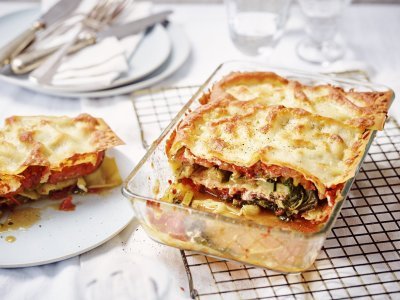 Vegetarische lasagne met snijbiet, courgette en ricotta De lasagne staat op tafel in een glazen doorzichtige ovenschotel. Hij is aangesneden zodat je mooi de laagjes ziet van pasta, groenten en saus. De portie die is uitgeschept ligt op een bordje er naast.