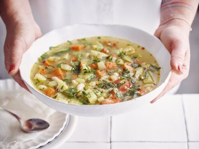 Een vegetarische minestrone met venkel, witte bonen, aardappel en verse kruiden geserveerd in een diep wit soepbord. Een vrouw met mooi rood gelakte nagels draagt het bord. 