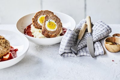 Vogelnestjes in tomatensaus met aardappelpuree In een diep wit bord ligt de aardappelpuree onderaan met verse rode tomatensaus en daar bovenop de doormidden gesneden vogelnestjes. Er ligt een handdoek langs met blauwe ruitjes, hierop het bestek met houten handvat.