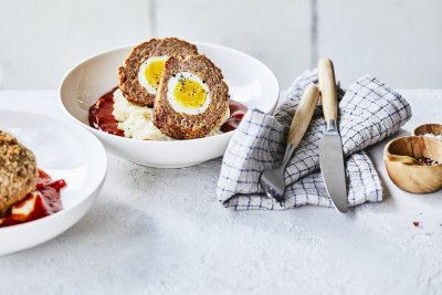 In een diep wit bord ligt de aardappelpuree onderaan met verse rode tomatensaus en daar bovenop de doormidden gesneden vogelnestjes. Er ligt een handdoek langs met blauwe ruitjes, hierop het bestek met houten handvat. 