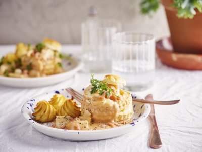 Vis vol-au-vent van schelvis en grijze garnalen Twee borden staan klaar om van te smullen. Vol-au-vent van vis in een bladerdeegpasteitje met pommes duchesse. Er staat een glaasje water naast het bord.