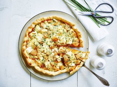 Zoete aardappeltaart met geitenkaas Quiche of groententaart van zoete aardappel met verse geitenkaas. Aangesneden op een wit bord met zwart randje en zilveren taartschep. Links van het bord staan witte molens van peper en zout en rechtsboven liggen enkele bieslooktakjes met keukenschaartje. Dit alles op een witte houten tafel.