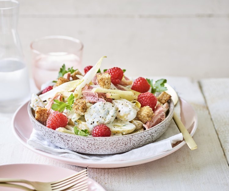Aardappelsalade met Breydelham, appel en framboos Een rijkelijke aardappelsalade is geserveerd in een grote kom. Je ziet de aardappel met de dressing, stukjes Breydelham, framboosjes en appel. De kom staat op een pastelkleurig roos bord. Er staat ook een karaf water langs met een glas in dezelfde tint als het roze bord.