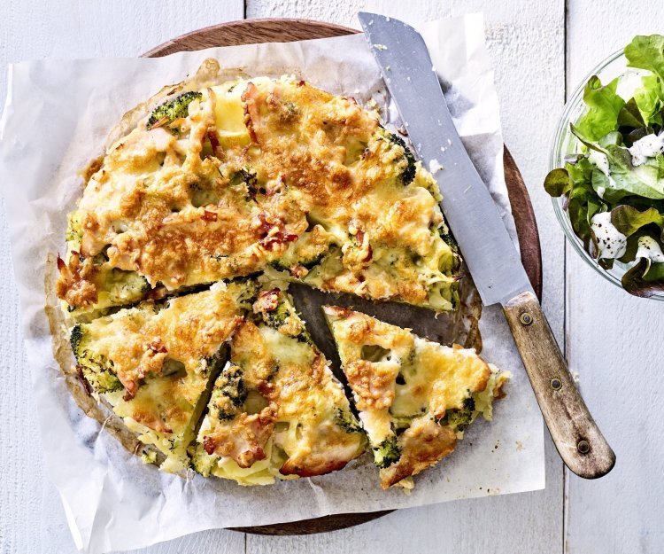 Deze hartige taart lijkt op een quiche maar is toch anders. Deze is gemaakt van aardappelschijfjes, broccoli en kippenwit. Ze ligt op een houten ronde snijplank met boterpapiertje onder, ze is al voor de helft aangesneden. Rechtsboven staat nog een kommetje gemengde sla. 