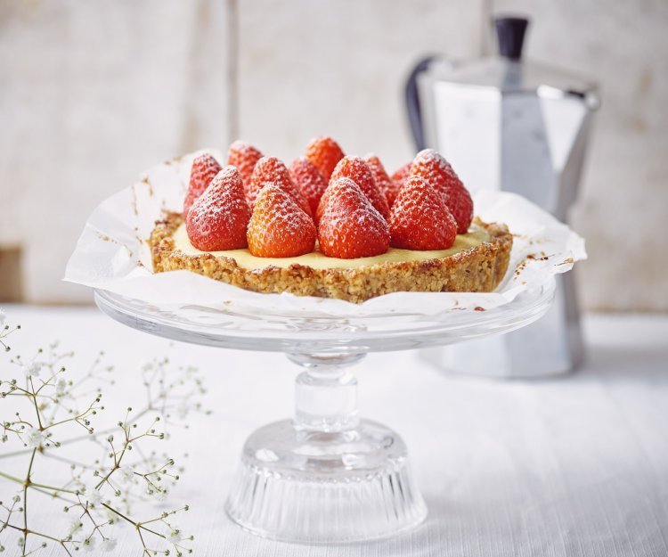 Aardbeientaart met vanillepudding en notenbodem De taart staat de stralen op een kristallen taartplateau. De verse aardbeien op de taart stelen de show. Achteraan de foto staat een kleine percolator om verse koffie te maken.