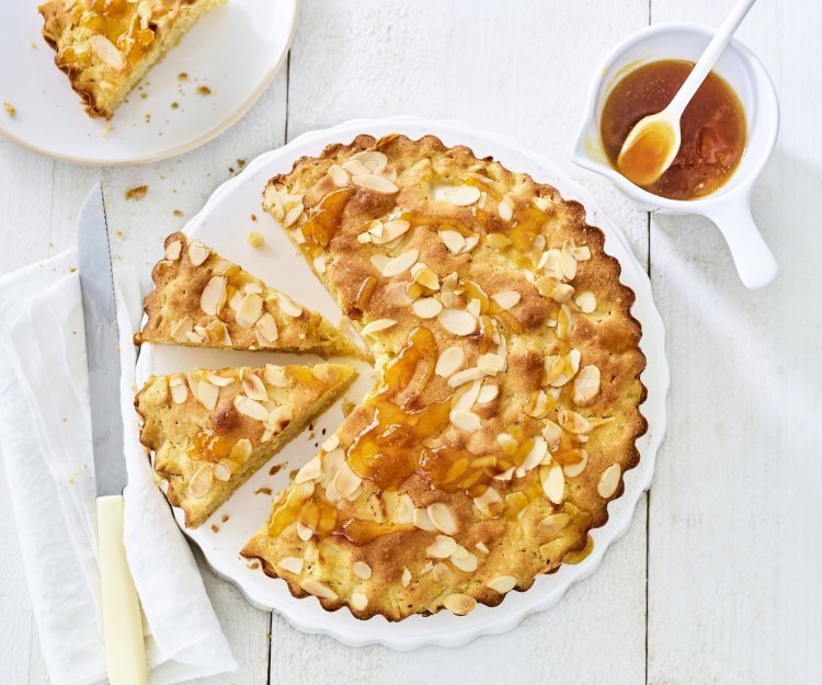 Goudgele appelcake staat op een houten witte tafel. De ronde cake is al voor een kwart aangesneden. Er is al een stukje uitgenomen en er staat een potje met abrikozengelei bij. 