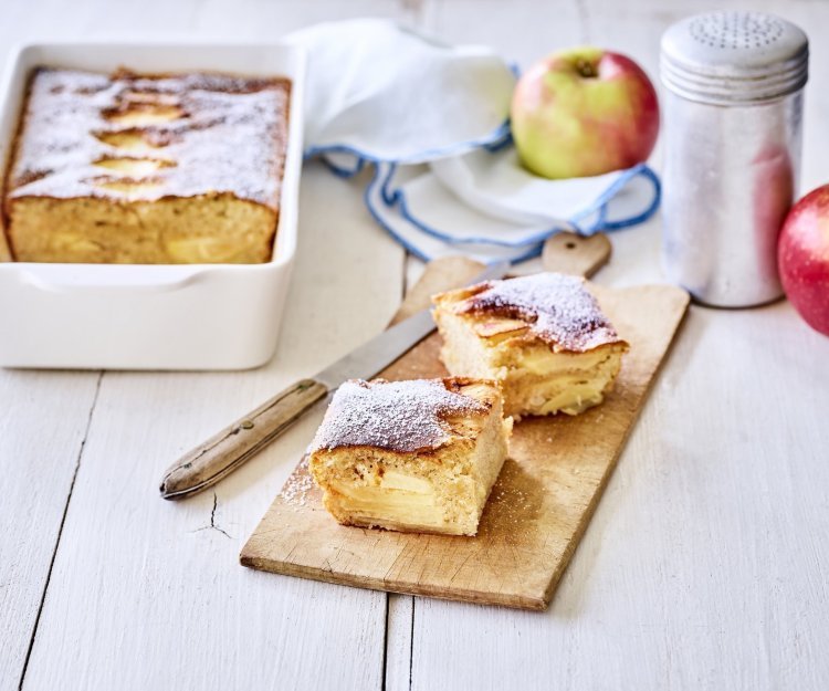 Appelcake van appelschijfjes Een verse appelcake uit de oven, je reukt hem waarschijnlijk al. Er is een stukje uitgesneden en deze ligt opnieuw aangesneden op een houten plankje. Op tafel ligt een witte servette met blauwe randjes, nog enkele verse appels en de strooipot met bloemsuiker.