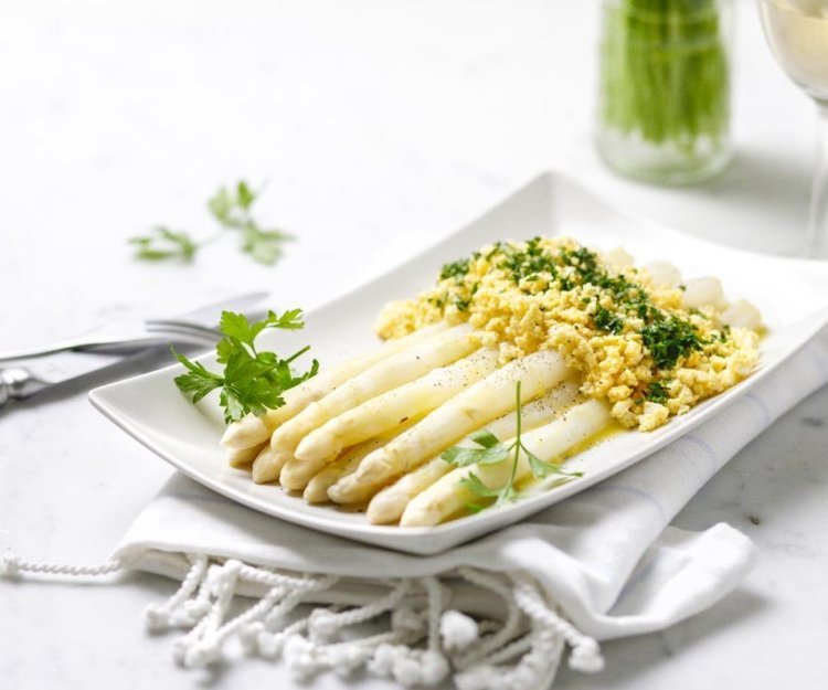 Op een rechthoekig wit bord liggen gekookte asperges, afgewerkt met geplette gekookte eieren, peterselie en gesmolten boter. Klaar om aan te vallen. 