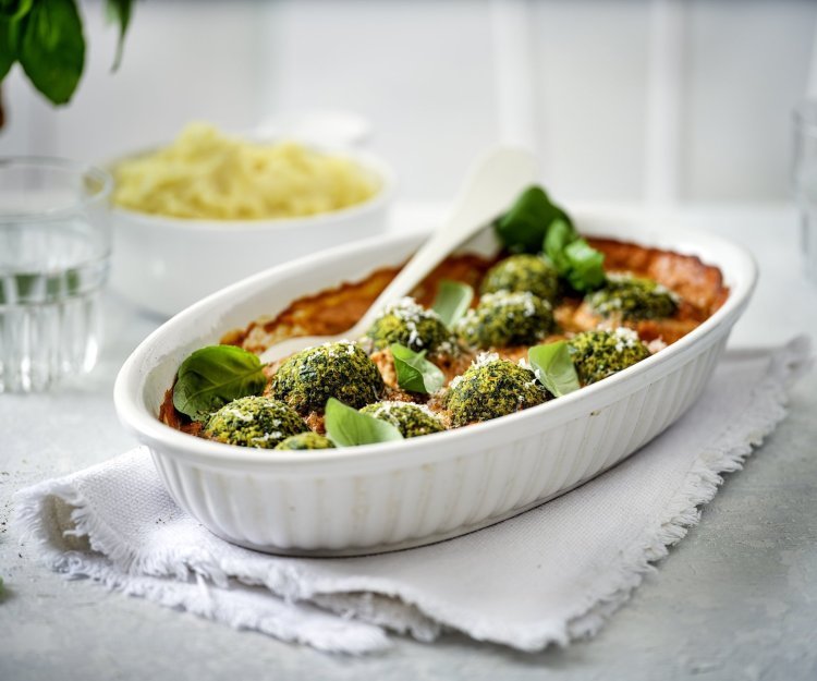 Balletjes van boerenkool in paprikasaus Een witte ovenschotel staat op tafel op een dikkere keukenhanddoek. De ovenschotel is gevuld met rode paprikasaus en hierop liggen de groene vegetarische balletjes van boerenkool. In de verte zie je ook nog een kommetje puree, klaar om aan tafel te gaan dus!