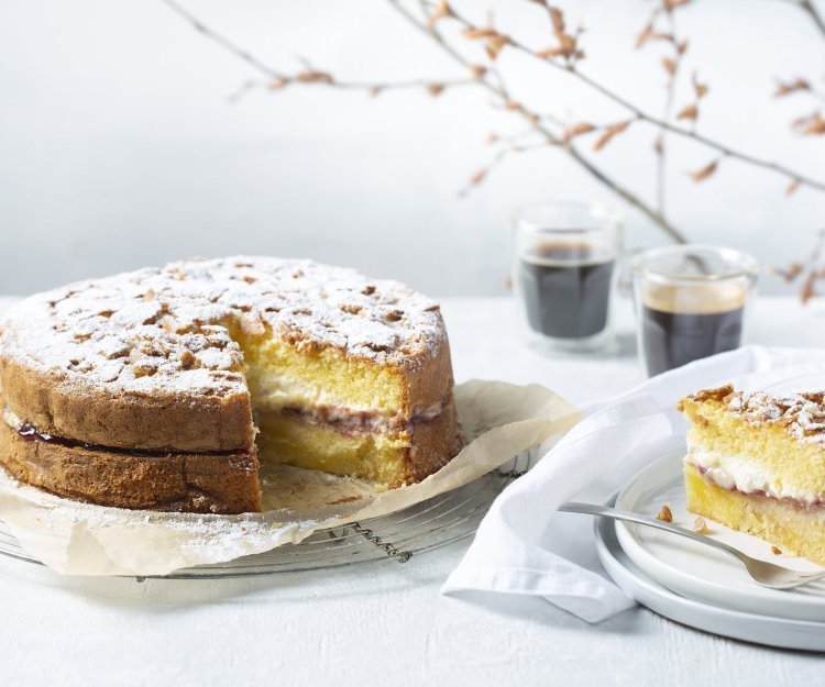 Een biscuit met vanillepudding en slagroom staat op tafel op een taartrooster. Er is al een stukje aangesneden om de lijnen duidelijk te laten zien van de pudding en slagroom. Koffie uitgeschonken, vorkje prikken en aanvallen die taart. 