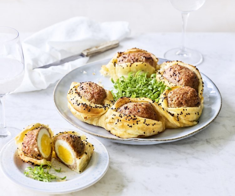 Bladerdeegkrans van vogelnestjes Op tafel staat een bord waarop een bladerdeegkrans ligt. Er zitten grote gehaktballen tussen gevluchten met hierin een gekookt eitje. Het bladerdeeg is afgewerkt met zwarte sesamzaadjes wat zorgt voor een leuke spikkeling.