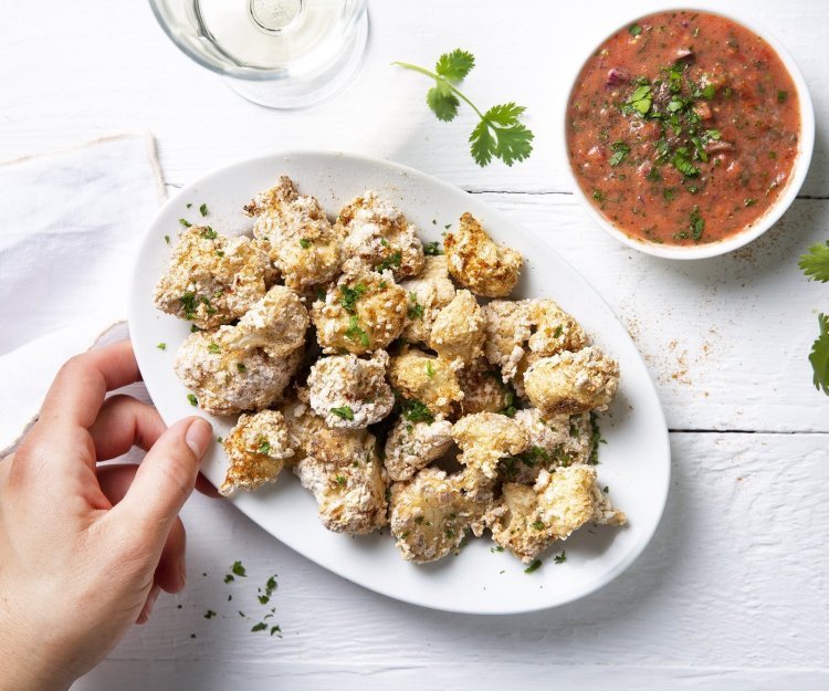 Bloemkoolbite met pittig sausje Krokante bloemkoolroosjes als aperitiefhapje, liggen klaar op een ovaal wit bordje. Er staat een pittige tomatensaus langs om te dippen. Tijd voor de apero te starten.