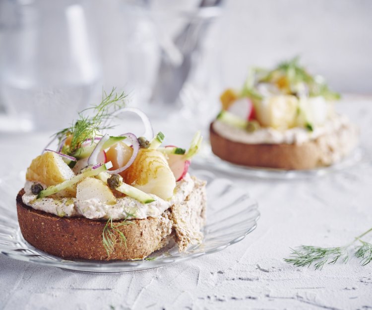 Bruschetta met bonenpuree, gerookte heilbot, radijzen en komkommer Een goedgevulde bruschetta ligt op een klein glazen bordje en staat op tafel. De bruschetta werd afgewerkt met een takje dille. Achteraan de tafel staat een kan met water en bestek om deze toast te verorberen.