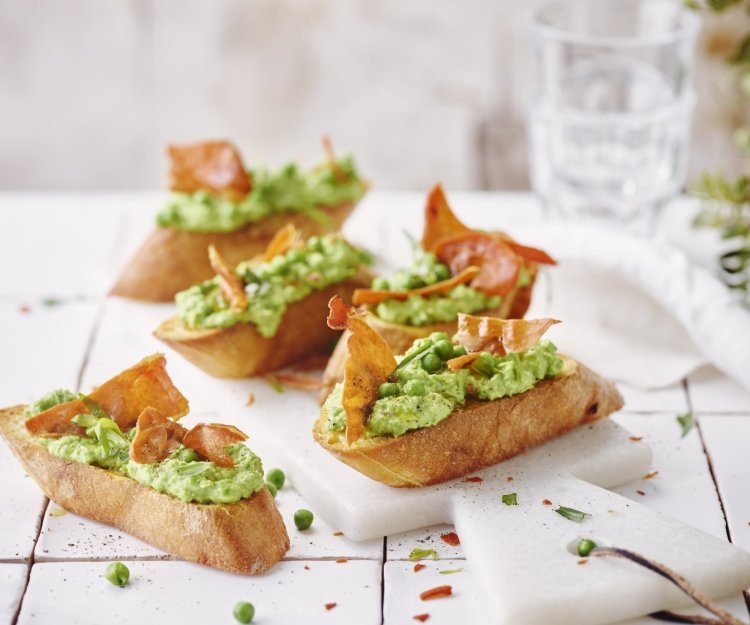 Bruschetta met geitenkaas, erwten, dragon en gedroogde ham Vijf klaargemaakte bruschetta liggen op tafel. De spread van geitenkaas en erwten kleurt mooi groen, de bruchetta werd afgewerkt met een stukje krokant gebakken gedroogde ham.