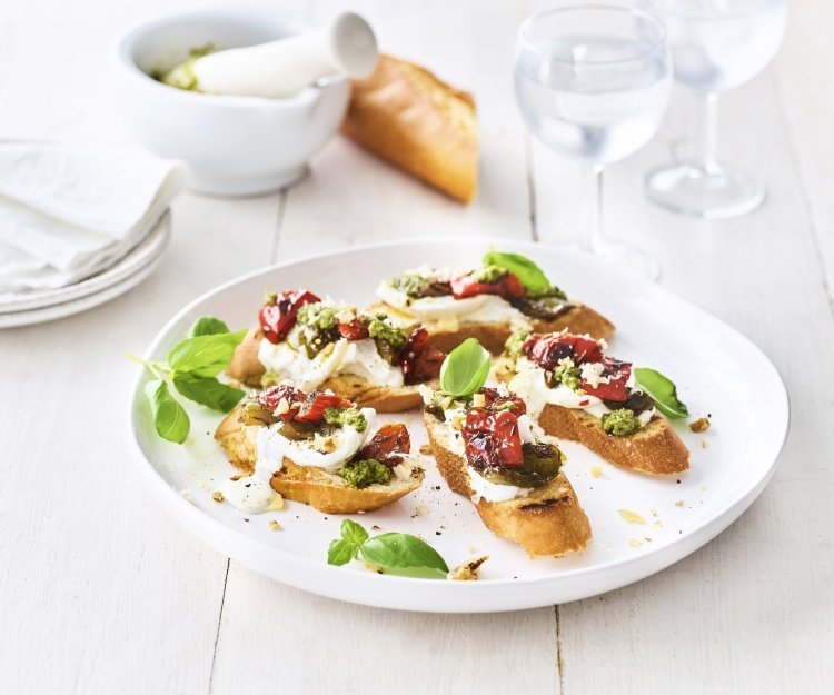 Bruschetta met paprika, burrata en pesto van basilicum met walnoten Op een houten, witte tafel staat een groot wit rond bord met hierop de bruschetta geserveerd. Ze zijn afgewerkt met extra blaadjes basilicum. Er staan ook kleine bordjes naast en de vijzel waarin de pesto is in gemaakt.