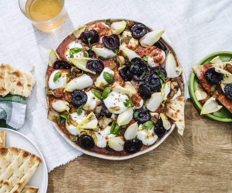 Burrata met gekaramelliseerd witloof en tomatenvinaigrette Op een houte tuintafel staat een grote ronde witte serveerschaal. Hij is gevuld met de bolletjes burrata, gebakken witlof en rode vinagrette op basis van tomaten. Wij serveren hem met krokant naanbrood en gebakken kersen.