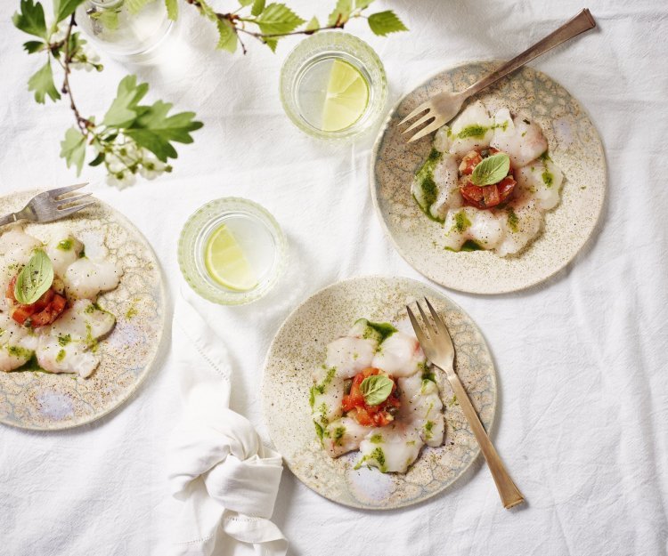 Carpaccio van zeeduivel met tomatentartaar en basilicumolie Drie feestelijke bordjes staan te schitteren op de tafel met een wit linnen tafelkleed. De vis is gesneden in fijne schijfjes en de tartaar van tomaten ligt in het midden van het bordje. Afgewerk met groene basilicum, heerlijke aperitiefhapje.