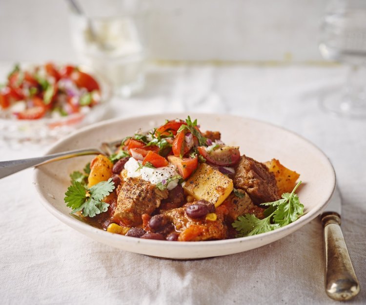 Chili con carne van stoofvlees met tomatensalsa en zure room Stoofvlees gemaakt met de smaken van een chili con carne, heerlijk! Er is een portie uitgeschept in een diep bord en staat op een gedekte tafel. In de achtergrond staat de tomatensalsa en zure room om het gerechtje helemaal af te werken.