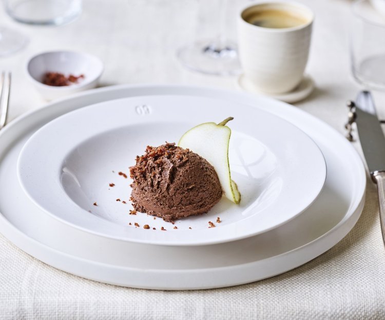 Chocomousse met een crumble van spek In een diep wit bord ligt een bolletje chocomousse met hierop de crumble van spek en een plakje peer. Bovenaan zie je een heerlijke kopje kofie staan. Perfect dessert om het feestmenu in glans af te sluiten.