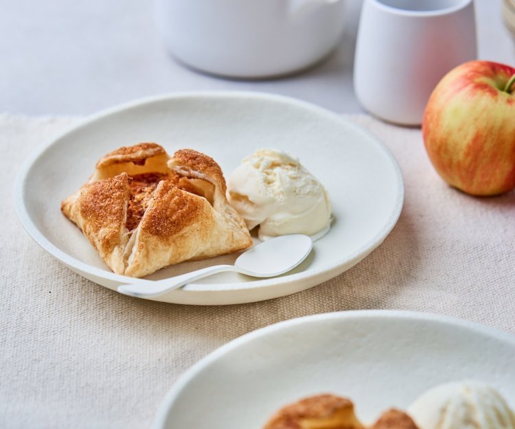 Op een mooi wit bord ligt een appeltaartje van bladerdeeg en een bolletje vanille-ijs. Er liggen ook enkele jonagold appelen langs het gerecht. 