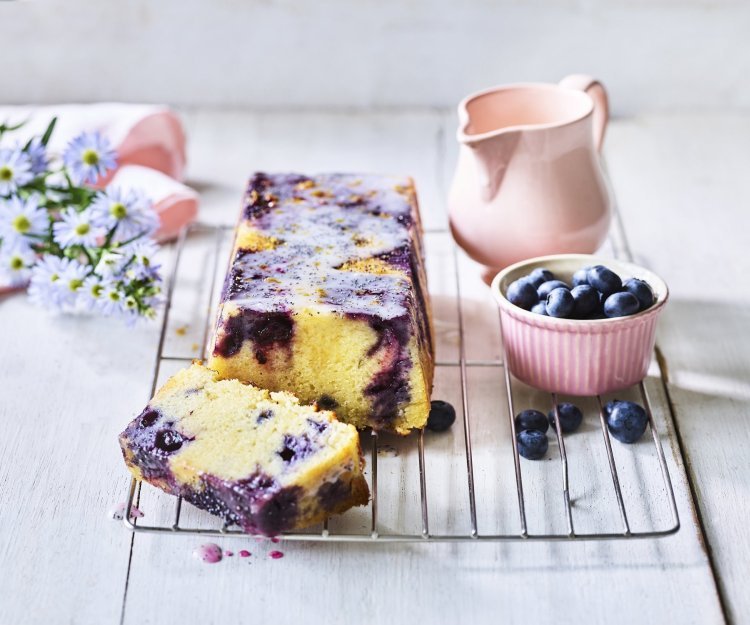 Citroencake met yoghurt, blauwe bessen en maanzaad Op een bakrooster ligt de citroencake af te koelen, hij heeft een prachtige kleur door de blauwe bessen die tinten van paars achterlieten in de smeuige cake. Er staat nog een extra portie blauwe bessen naast voor extra fruitplezier.