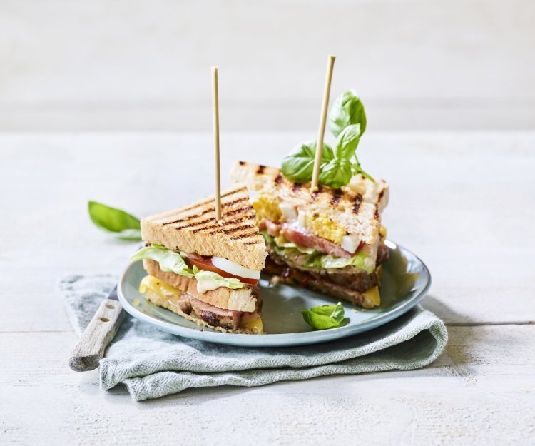 Club sandwich met gegrilde lamsfilet Goedgevulde club sandwich met lamsvlees en rauwkost. Gepresenteerd op een bord met middenin een prikker, klaar om op te eten.