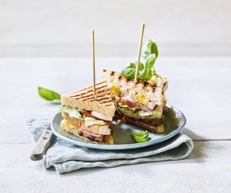 Goedgevulde club sandwich met lamsvlees en rauwkost. Gepresenteerd op een bord met middenin een prikker, klaar om op te eten. 