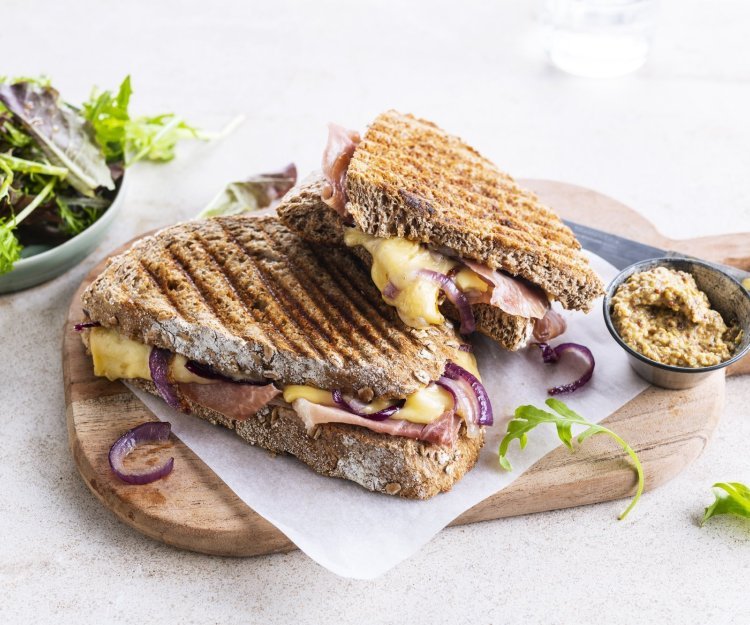 Croque met ham, rode ui en Passendale kaas De croque ligt op een houten plankje aangesneden. De kaas en rode ui zien er heerlijk uit. In de achtergrond zie je ook nog wat gemengde sla en een potje graantjesmosterd.