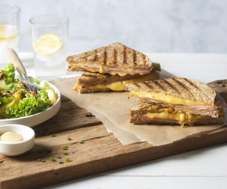 Een krokant gebakken croque monsieur ligt aangesneden op een houten snijplank. Lang staat een salade in een diep wit bord met een potje mayonaise voor te dippen. 