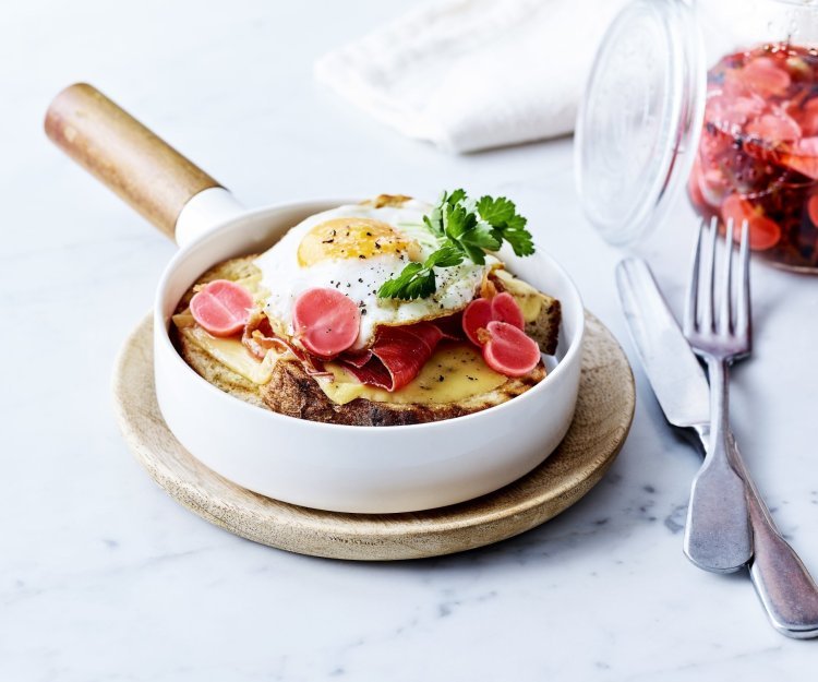 Croute au fromage met Averbode kaas in een witte pan, spiegeleitje er bovenop gepresenteerd met een takje verse peterselie. Op de achtergrond staan gepekelde radijzen in glazen pot.