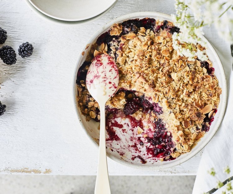 Crumble met havermout en braambessen Crumble met havermout en braambessen in een witte ronde ovenschaal. Er is al een portie crumble verdwenen uit de ovenschaal en er liggen enkele braambessen langs. Om nog wat extra sfeer toe te voegen zijn er witte fijne bloemetjes rechts in beeld.
