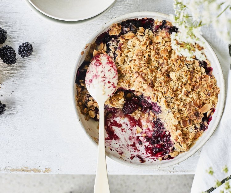 Crumble met havermout en braambessen in een witte ronde ovenschaal. Er is al een portie crumble verdwenen uit de ovenschaal en er liggen enkele braambessen langs. Om nog wat extra sfeer toe te voegen zijn er witte fijne bloemetjes rechts in beeld.