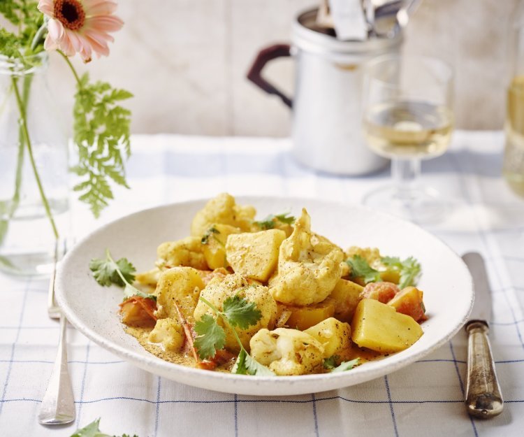 Heerlijke goudkleurige curry geserveerd in een diep wit bord met bloemkool en aardappelen. Er ligt bestek langsheen het bord en glaasje witte wijn. Ook zien we een vaasje met roze bloem en varentakje. 