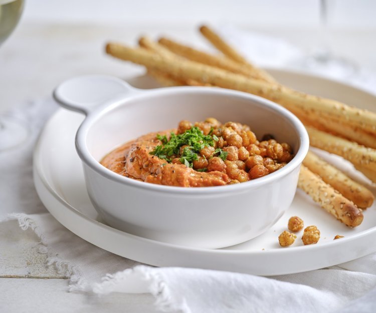 Dip van zoete puntpaprika met geroosterde kikkererwten Een wit porseleine potje is gevuld met een oranjekleurige tapenade. Het is er eentje van zoete puntpaprika met hierop geroosterde kikkererwten. Er liggen broodstengels langs om te dippen.