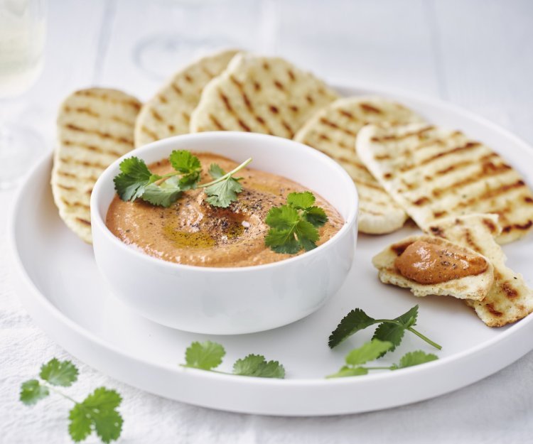 Dip van zoete puntpaprika met naanbrood Homemade paprikadip zit in een wit schaaltjes. Het naanbrood ligt er langs op een wit bord. Het gercht is afgewerkt met korianderblaadjes.
