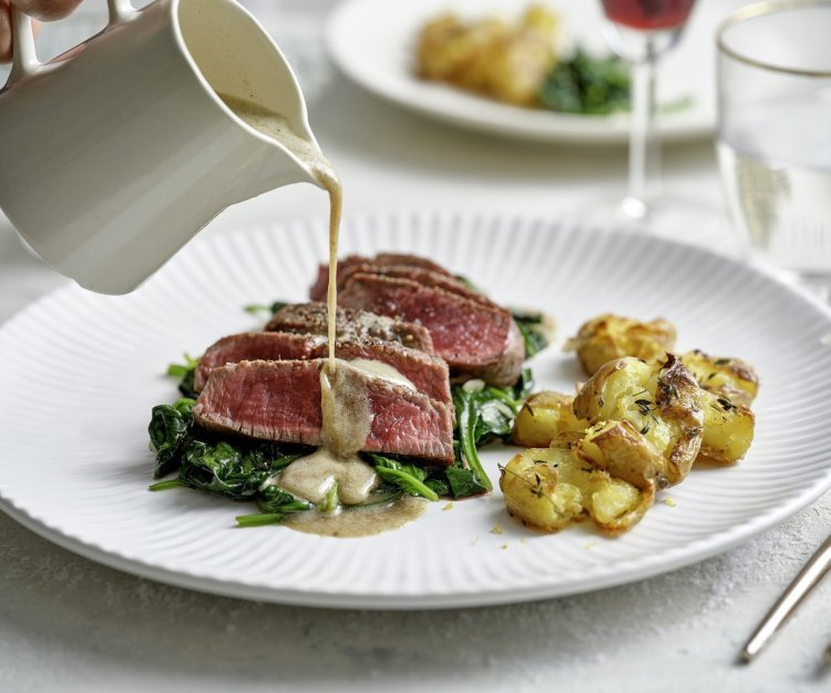 Filet pur met blauwe kaas en gecrushte aardappelen Een wit bord staat op tafel met hierop de heerlijke krokante aardappel en de saignant gebakken filet pur op een bedje van spinazie. Er komt een sauskannetje in beeld die saus over het vlees giet. De tafel is gedekt voor twee met de perfecte maaltijd.